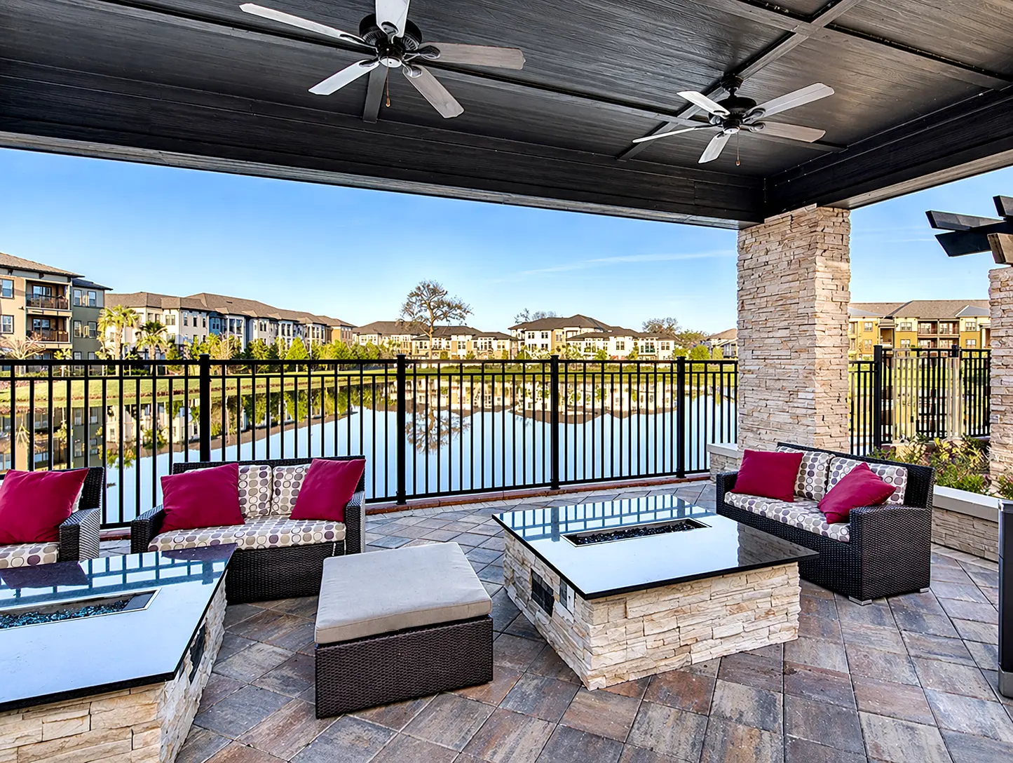 A stylish outdoor lounge area featuring dark wicker furniture with colorful cushions, set under a shaded patio with ceiling fans. The backdrop includes a serene view of a lake and residential buildings.
