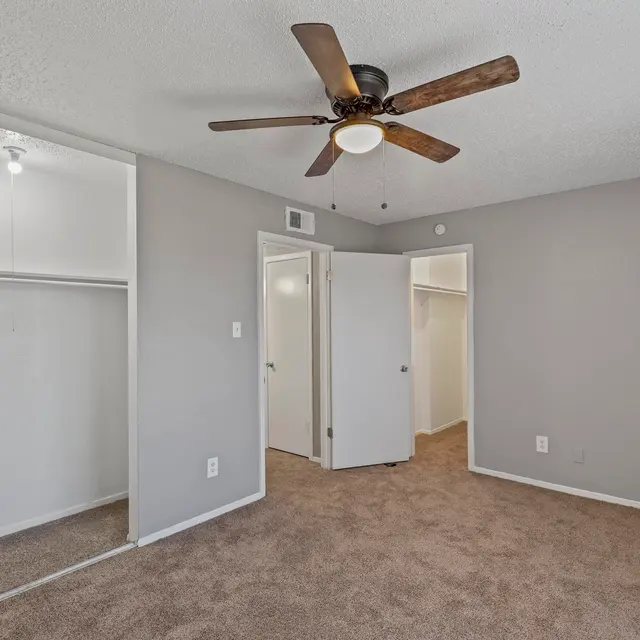 A spacious bedroom featuring beige carpeting, light gray walls, and a ceiling fan. There's a large closet on the left and doors leading to other rooms on the right.