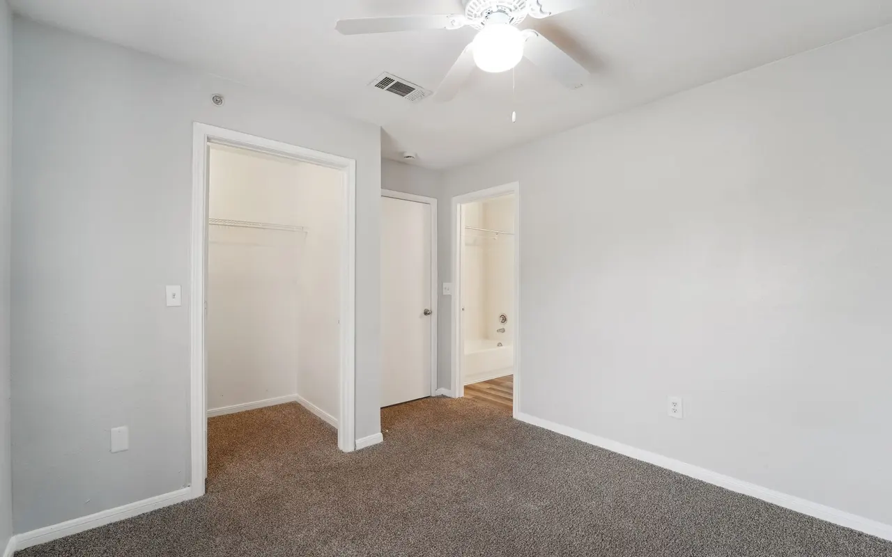 A light-colored room featuring carpeted flooring and a ceiling fan. Two doors are visible—one leading to a closet and the other to a bathroom. The walls are painted in a neutral tone, contributing to a spacious feel.