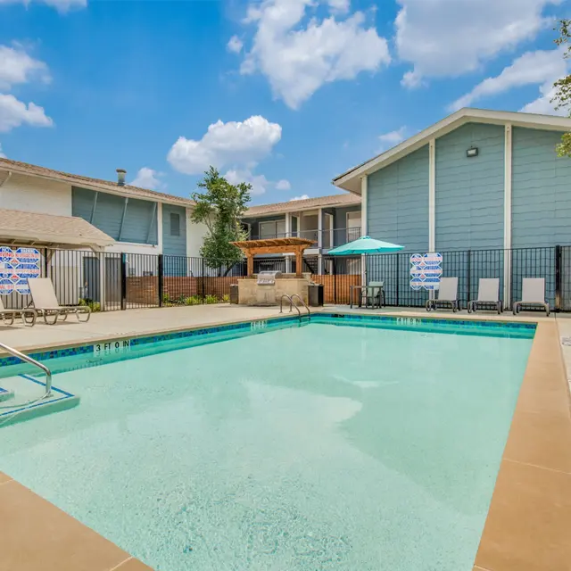 A swimming pool area in an apartment complex with lounge chairs and an umbrella in the background. The sky is partly cloudy.