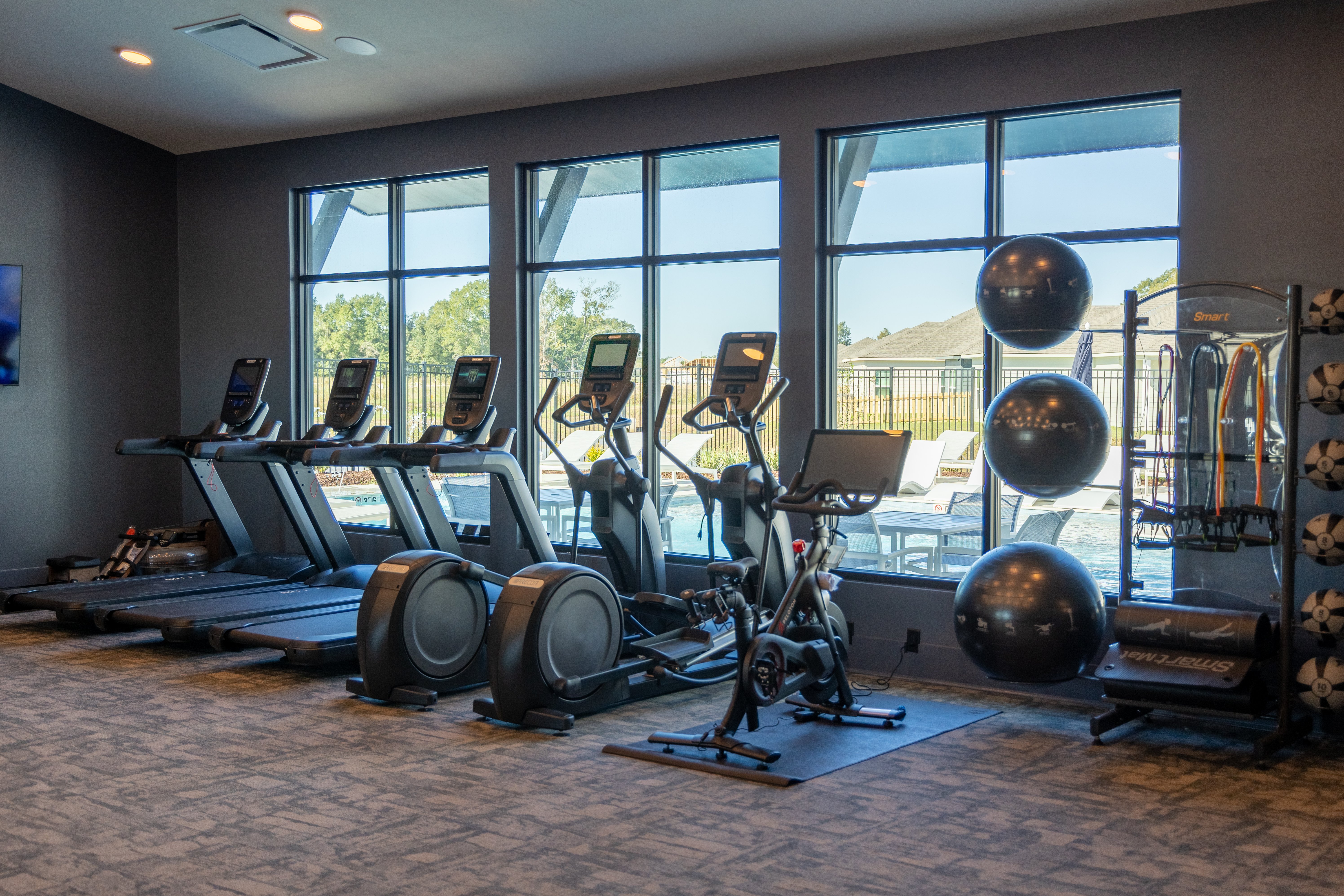 An indoor gym area featuring several treadmills, elliptical machines, and a stationary bike next to a wall with large windows. In the background are exercise equipment and fitness balls.