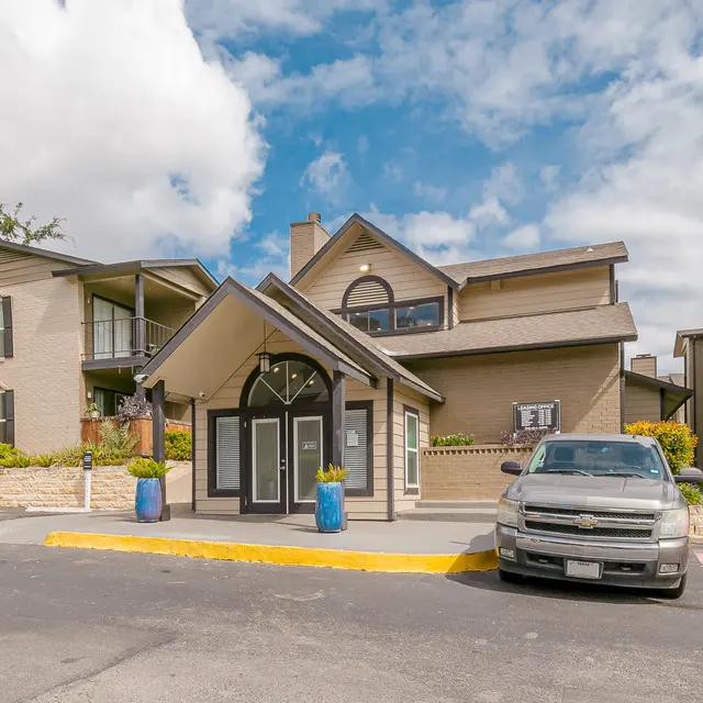 Entrance of an apartment complex with a modern architectural design, featuring two-story buildings, parking area, and blue planters.