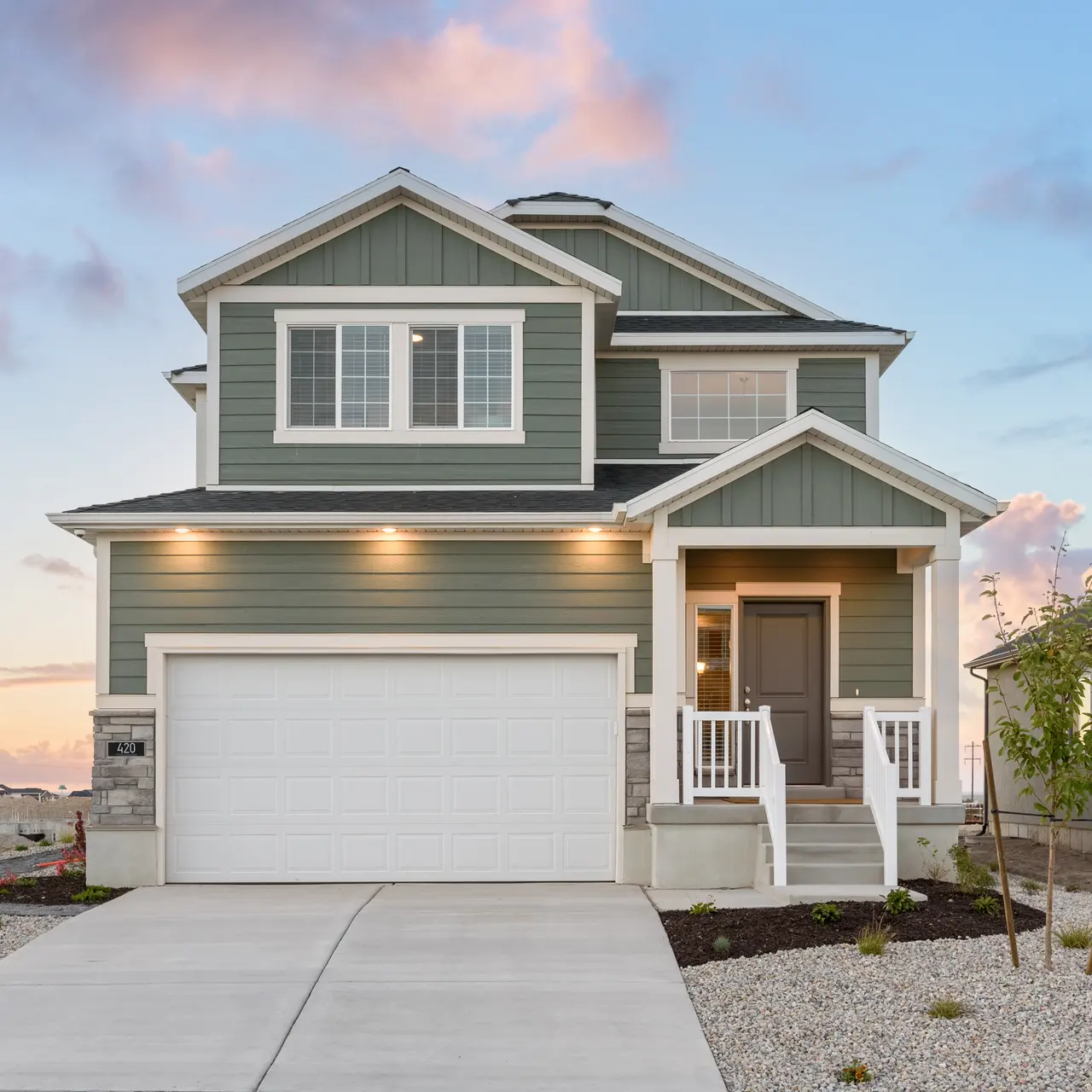 A modern two-story house with green siding and a white garage door, set against a sunset sky. The house features a front porch with white railings and landscaped gravel yard.