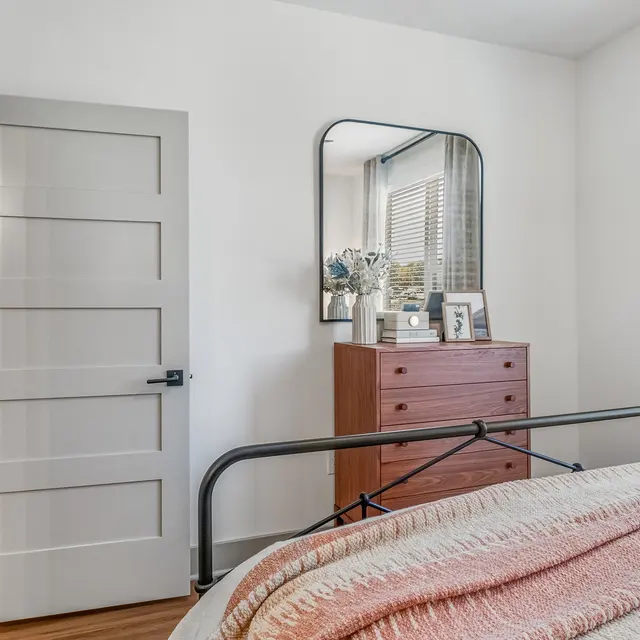 Cozy Modern Bedroom Interior A modern bedroom featuring a metal bed frame with a pink and white blanket, a wooden dresser with decorative items, a mirror on the wall, and a partially open door.