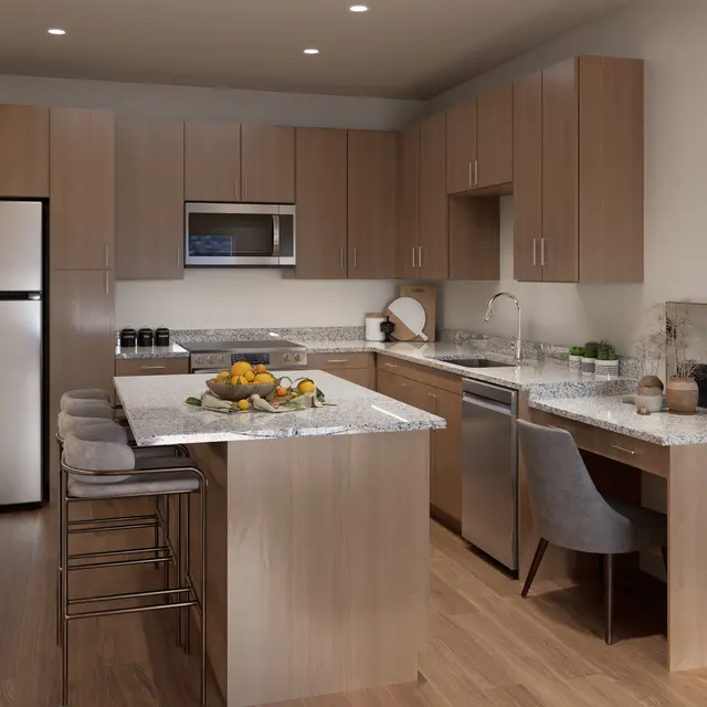 A modern kitchen featuring light wood cabinetry, granite countertops, and stainless steel appliances. There is a kitchen island with a bowl of fruits on it, bar stools, and an adjacent desk area.