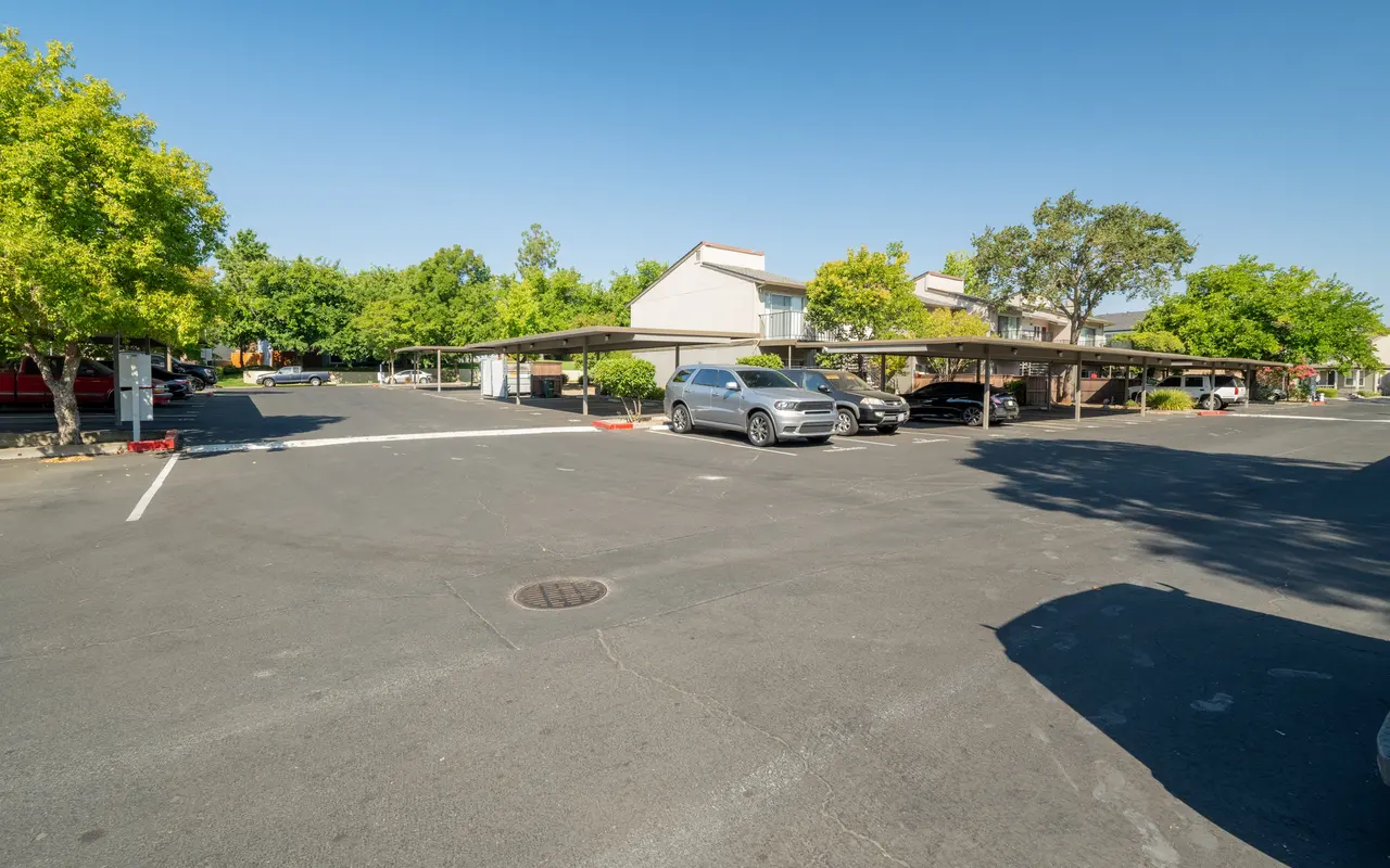 A view of a wide parking lot under a clear blue sky, featuring shaded carports and trees around the area.