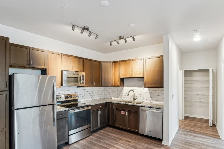 A modern kitchen featuring stainless steel appliances, wooden cabinets, and a tile backsplash. The kitchen includes a refrigerator, microwave, dishwasher, and a double sink, with ample counter space and polished wooden flooring.
