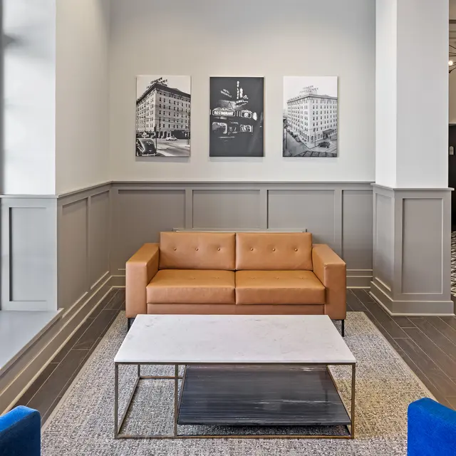 A modern living room featuring a tan leather couch, blue armchairs, and a coffee table on a patterned rug. The wall is adorned with three black and white photographs.