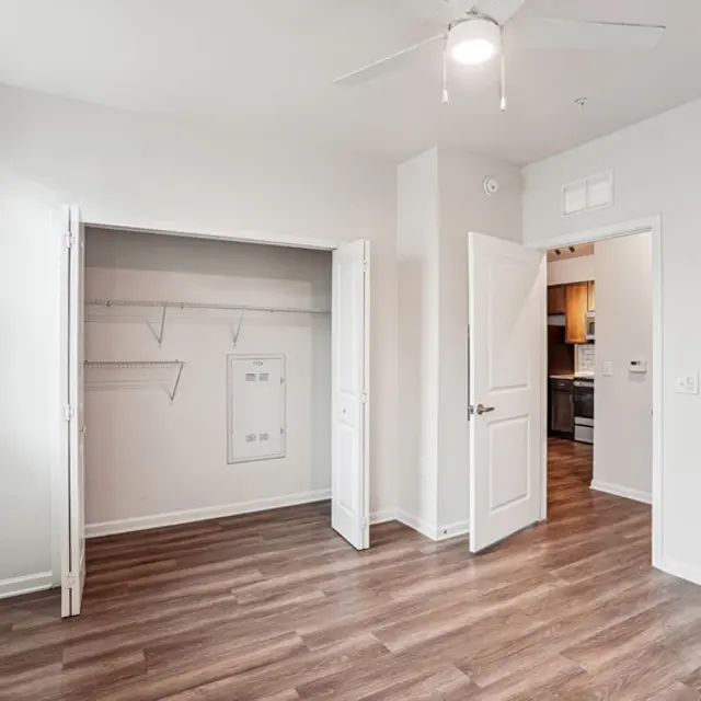 Empty room with light-colored walls, a ceiling fan, a closet with open doors, and a door leading to a kitchen area.