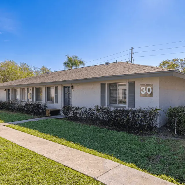 Apartment Building Exterior 30 A single-story apartment building with several windows, a green lawn in front, and a pathway leading to the entrance. The building has a number '30' displayed on the wall.
