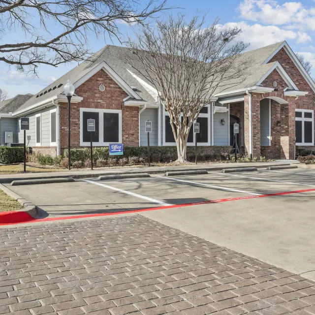 A brick building with a grey roof labeled 'Information Center' in front. It features large windows, a well-maintained lawn, and a paved parking area with a smooth surface. In the background, there are trees and a blue sky with white clouds.