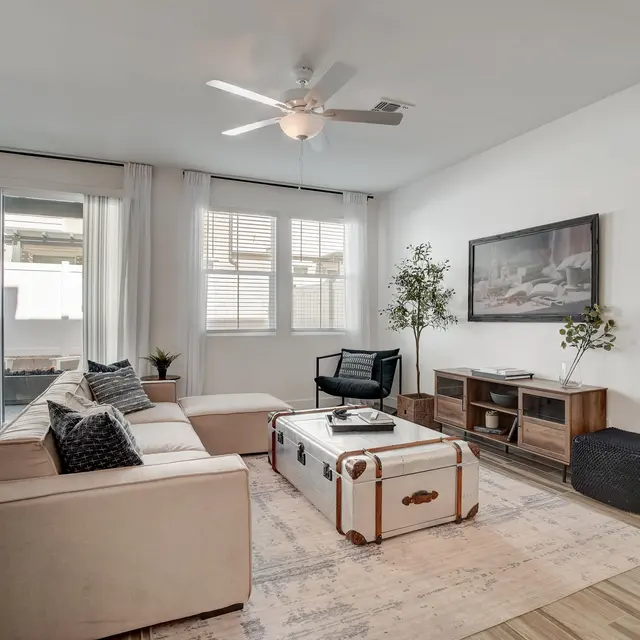 Modern Living Room Interior A stylish living room featuring a light-colored sectional sofa, a decorative coffee table, and a television stand with a TV. The room has a large window with sheer curtains, allowing natural light in. Two plants add a touch of greenery, while a cozy rug sits on the wooden floor.