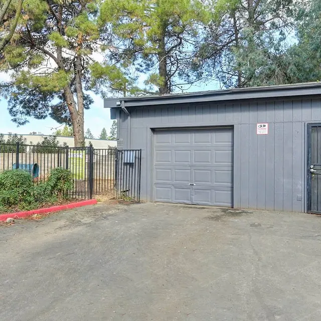 A gray storage building with a roll-up door and a side entrance, surrounded by trees and a fence.