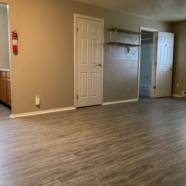 A spacious room with light brown laminate flooring, two closed doors on the right, a window providing natural light, and a small kitchenette with wooden cabinets visible on the left.
