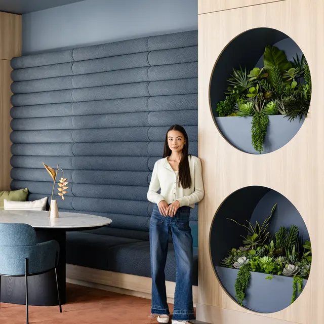 A young woman stands beside a decorative wall featuring circular planters filled with greenery. She wears a light blouse and jeans, and the setting includes a stylish seating area with a round table and upholstered chairs against a textured wall.