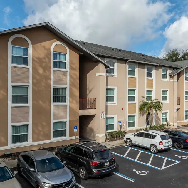 A multi-story apartment complex with beige exterior. The building features several large windows and a small balcony. A parking lot in front has multiple parked cars, and there are palm trees and blue skies in the background.