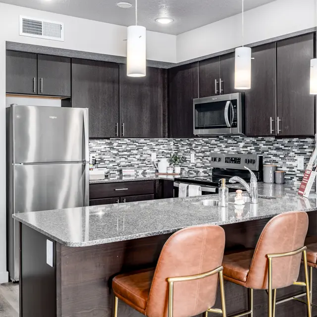 Modern Kitchen Design A modern kitchen featuring dark cabinetry, stainless steel appliances, and a stylish tile backsplash. The kitchen island has four brown leather bar stools, and there are pendant lights hanging above.