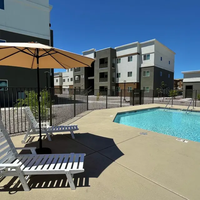 Pool Area at Apartment Complex A sunny pool area with two white lounge chairs and a large umbrella beside a turquoise swimming pool, surrounded by a black fence and modern apartment buildings in the background.