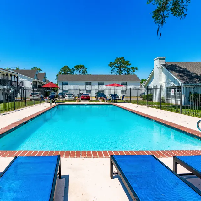 A bright blue swimming pool surrounded by lounge chairs and greenery, with a fenced area and buildings in the background.