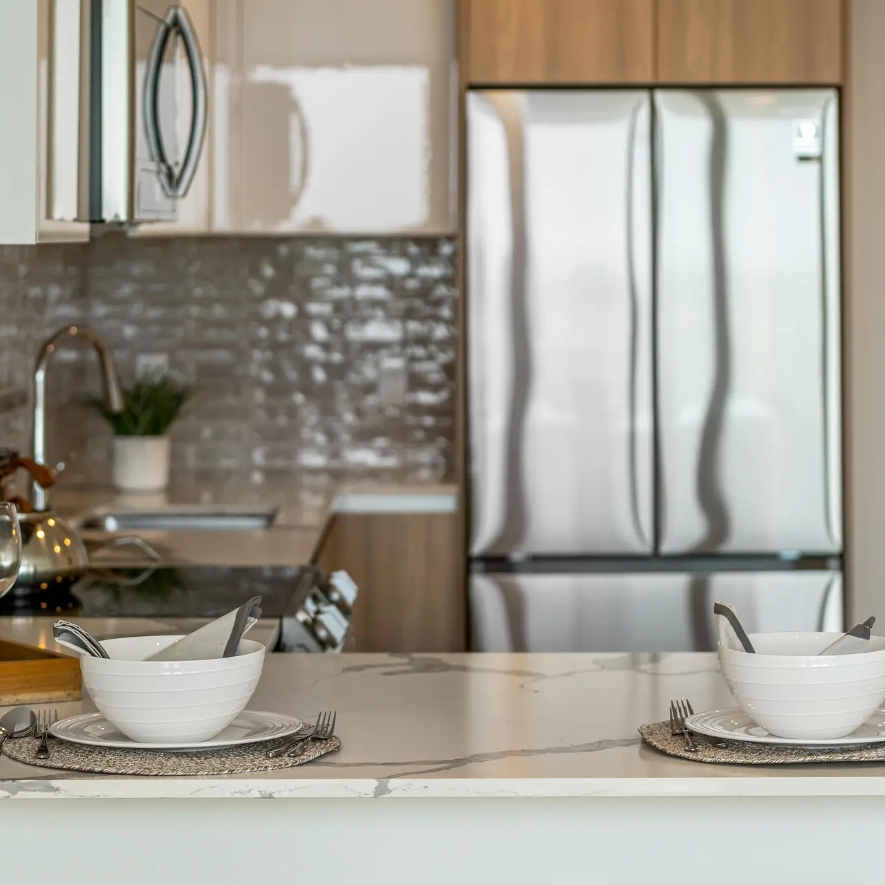 A modern kitchen with a sleek countertop featuring two bowls, utensils, and drinkware. Behind is a shiny silver refrigerator and a stylish backsplash.