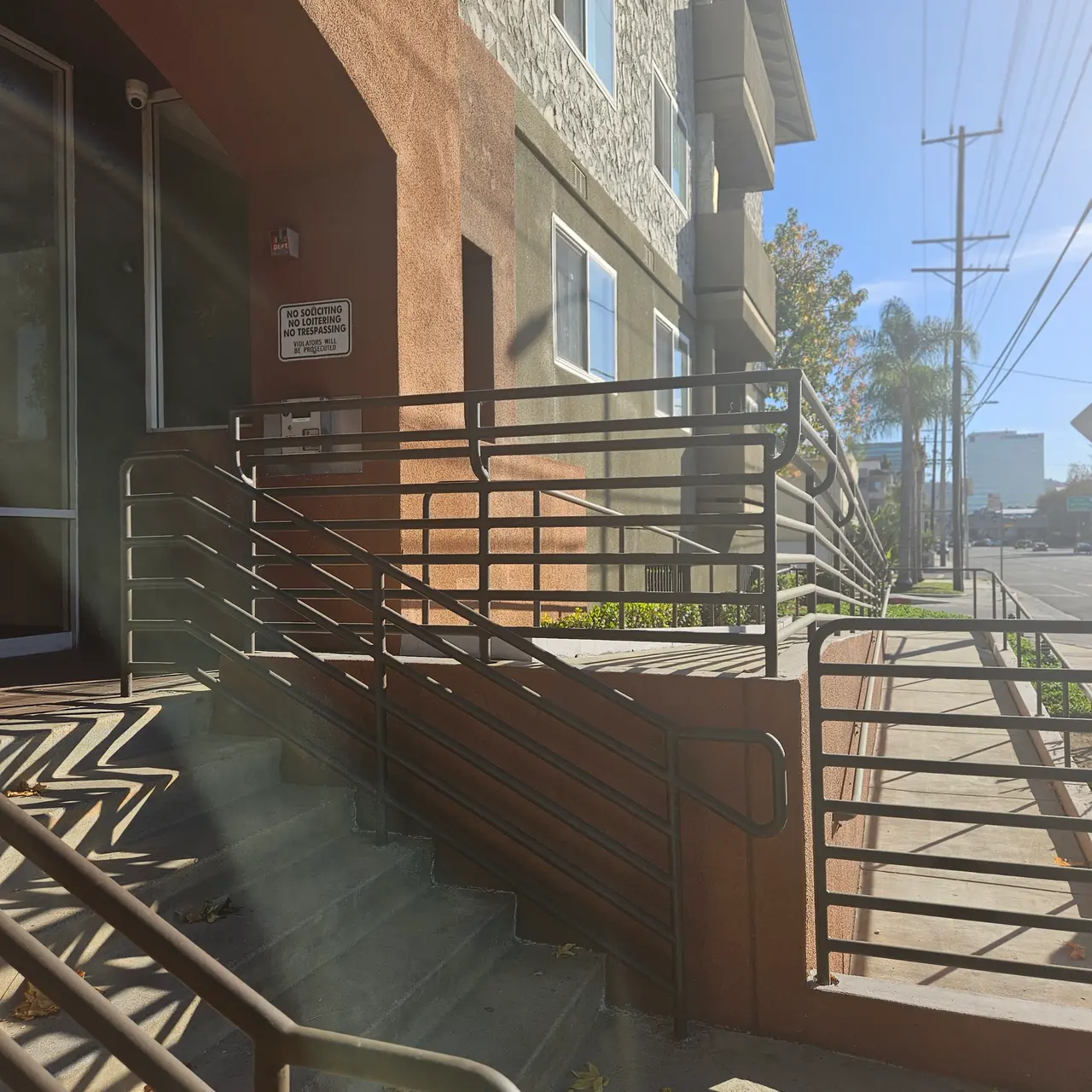 A modern building exterior showing a wheelchair accessible ramp with railings leading to the entrance. The ground is sunny with shadows cast by the railings and the building. Power lines are visible in the background.