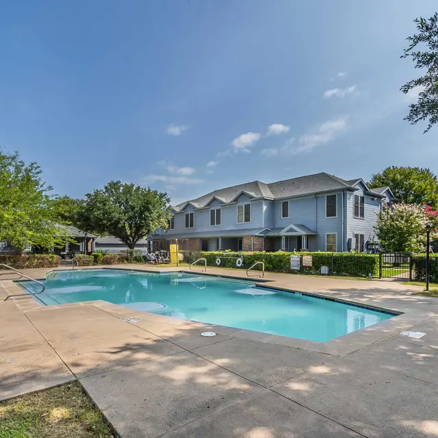 A community pool area featuring a clear blue swimming pool surrounded by pavement, green shrubs, and trees under a bright sky. In the background, there are residential buildings with outdoor seating areas.