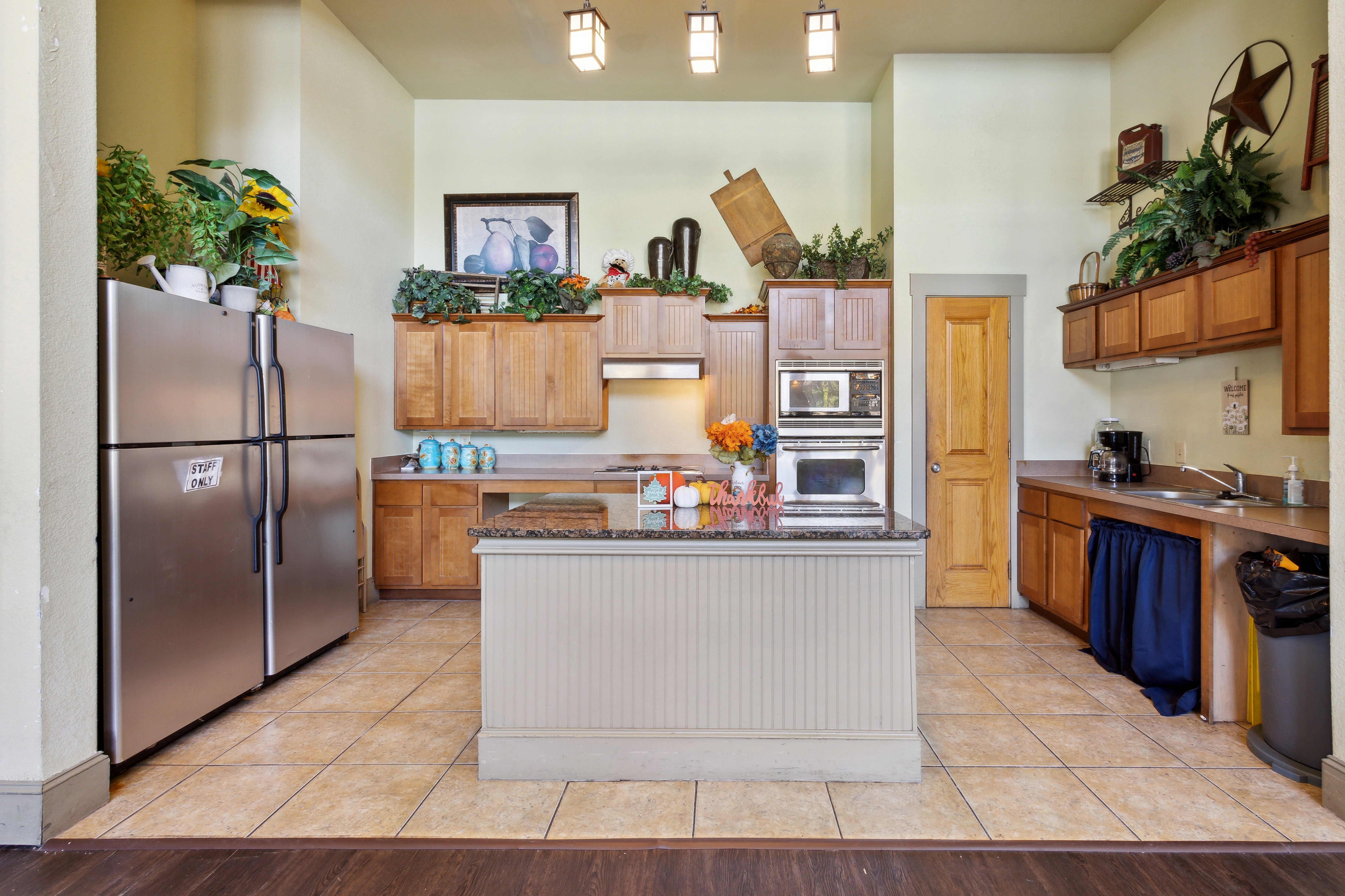 Spacious Modern Kitchen A spacious kitchen with wooden cabinetry, modern appliances, and a central island. Light-colored walls and tile flooring create a bright ambiance. Decorative items and plants add a homey touch.