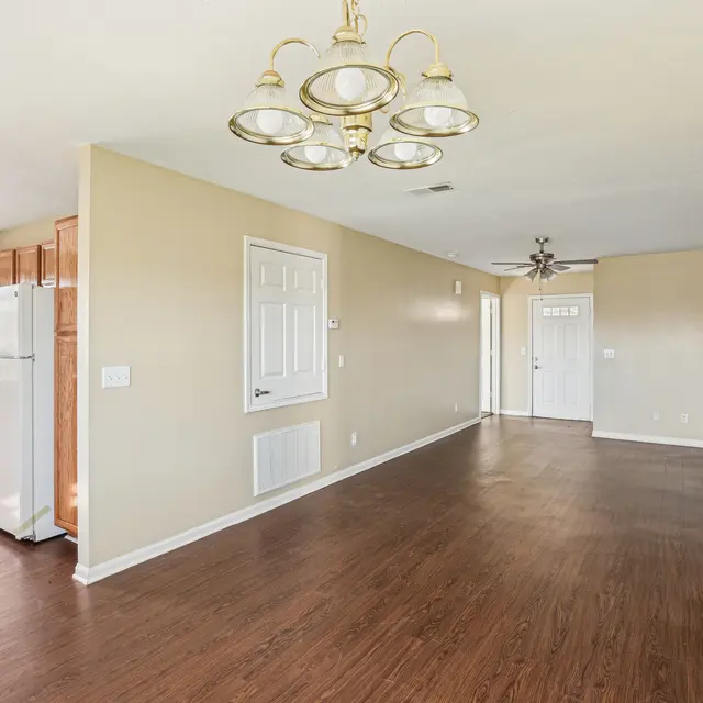 A spacious living area with hardwood flooring, beige walls, and a ceiling light fixture. There are kitchen cabinets on the left, with a refrigerator and a doorway leading to another room. Natural light is coming in through a window.