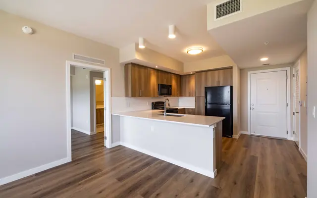 A modern apartment kitchen with sleek wooden cabinets, a black refrigerator, and a spacious island. The floor is made of light-colored wood, and there are large windows allowing natural light to enter.
