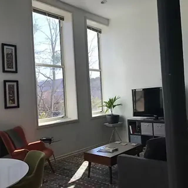 A modern living room with large windows letting in natural light. There are framed artworks on the wall, a television on a stand, a small coffee table, and a stylish grey couch. A plant is situated in the corner by the window, and there's a round dining table with colorful chairs in the foreground.