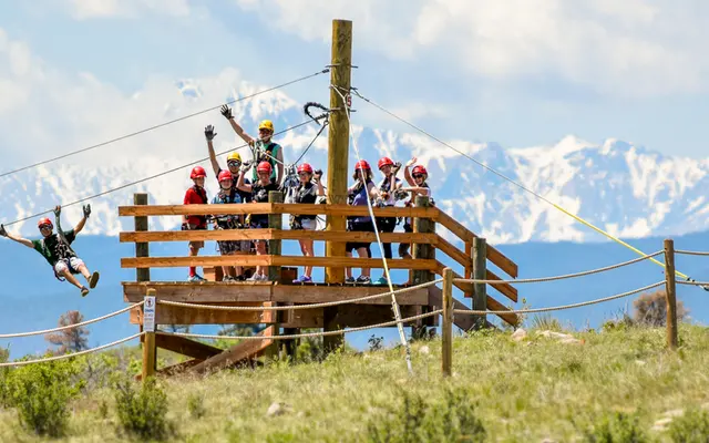 A group of thrill-seekers waiting on a wooden platform while one person zips down a zip line against a backdrop of snow-capped mountains.