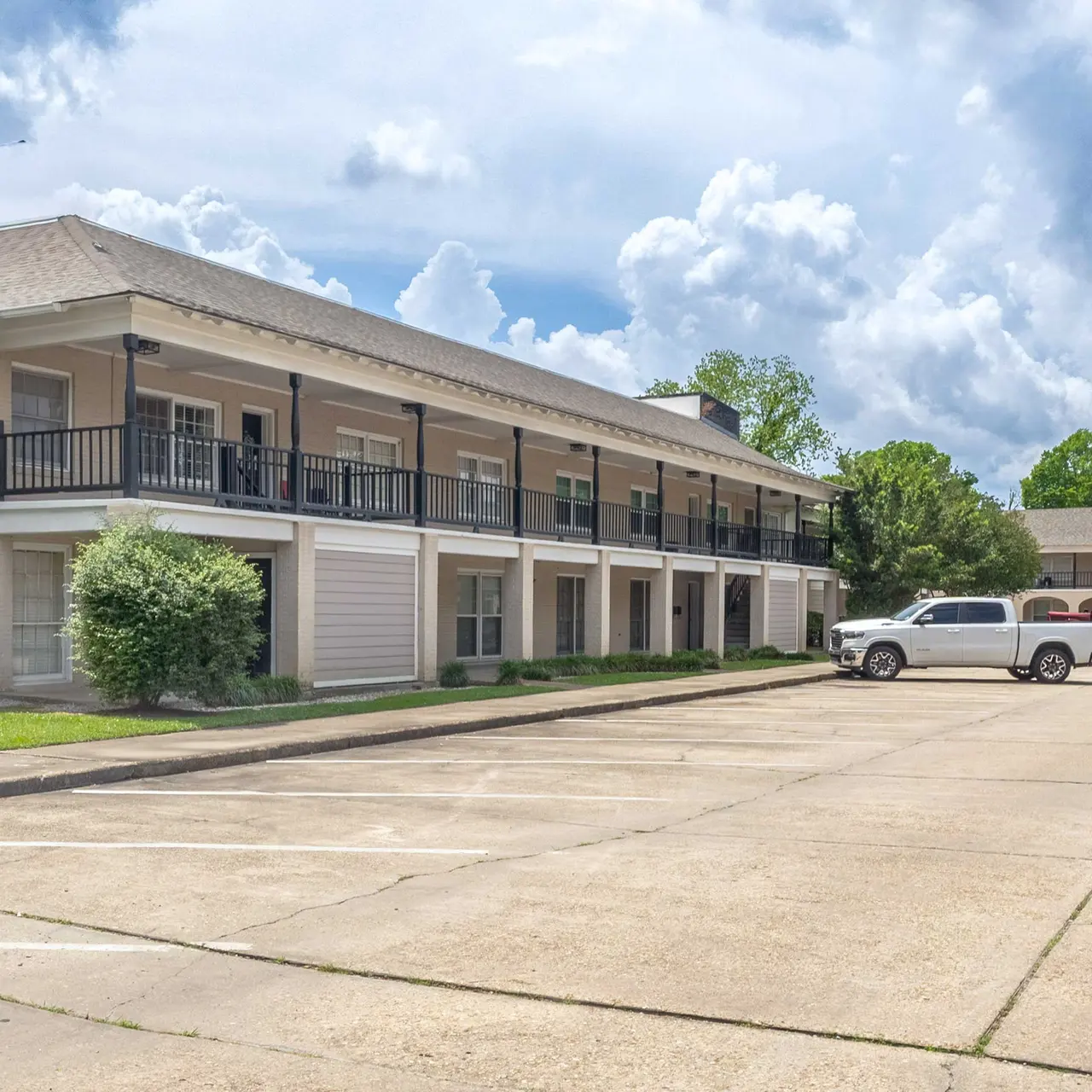 Exterior view of a multi-unit apartment building with two levels, featuring a parking lot in front and green grass around.