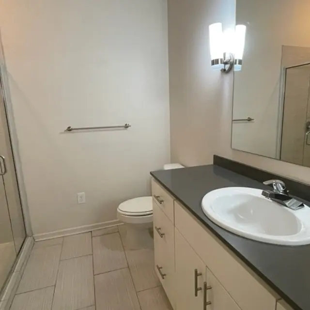A modern bathroom featuring a glass shower, a white toilet, and a sleek countertop with a sink. The walls are light-colored and there is soft lighting from a wall fixture.