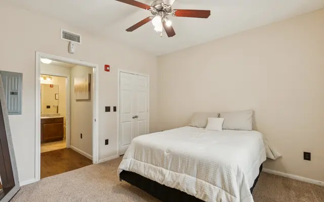 Bright and Spacious Bedroom A light-colored bedroom featuring a double bed with a white bedspread, a ceiling fan with wooden blades, and beige walls. There is a doorway leading to a bathroom visible in the background, and the floor is carpeted.