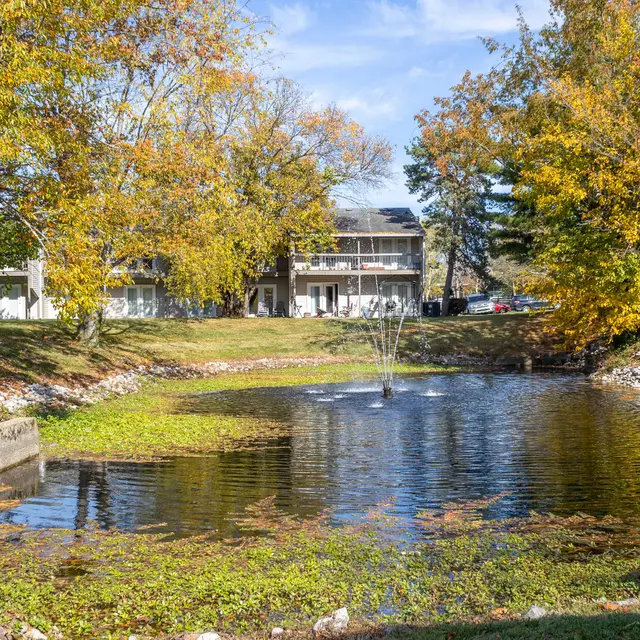 Autumn Pond View A serene pond surrounded by trees with autumn leaves and a small fountain in the center. Apartment buildings are visible in the background.