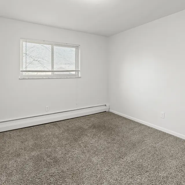 A vacant room with beige carpet and white walls, featuring a small window and door on the left.