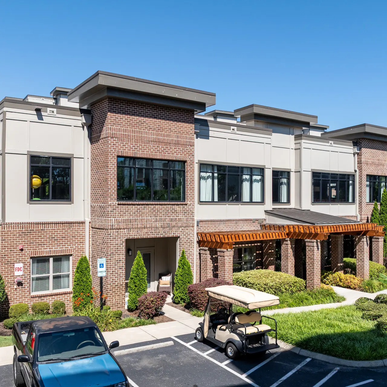 Modern apartment building with brick facade and large windows, surrounded by lush landscaping and parking area.
