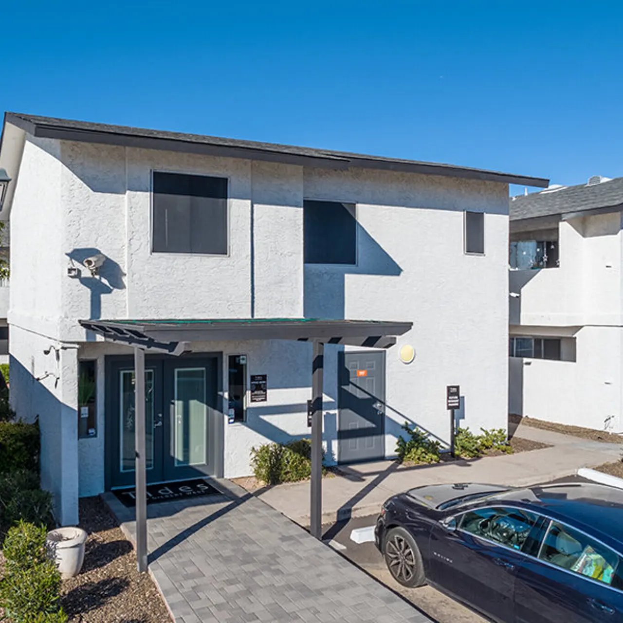 Exterior of a two-story apartment building with white walls and a modern design. The entrance features a covered porch, and there are parked cars nearby.