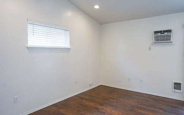 Empty Room Interior An empty room with white walls, a wooden floor, and a small window with blinds. An air conditioning unit is mounted on the wall.