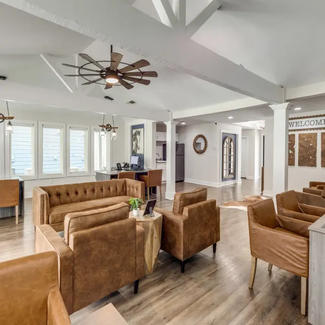 A spacious and inviting lobby area featuring brown leather sofas and chairs, wooden flooring, and large windows. There's a welcome sign on the wall and a reception desk in the corner.