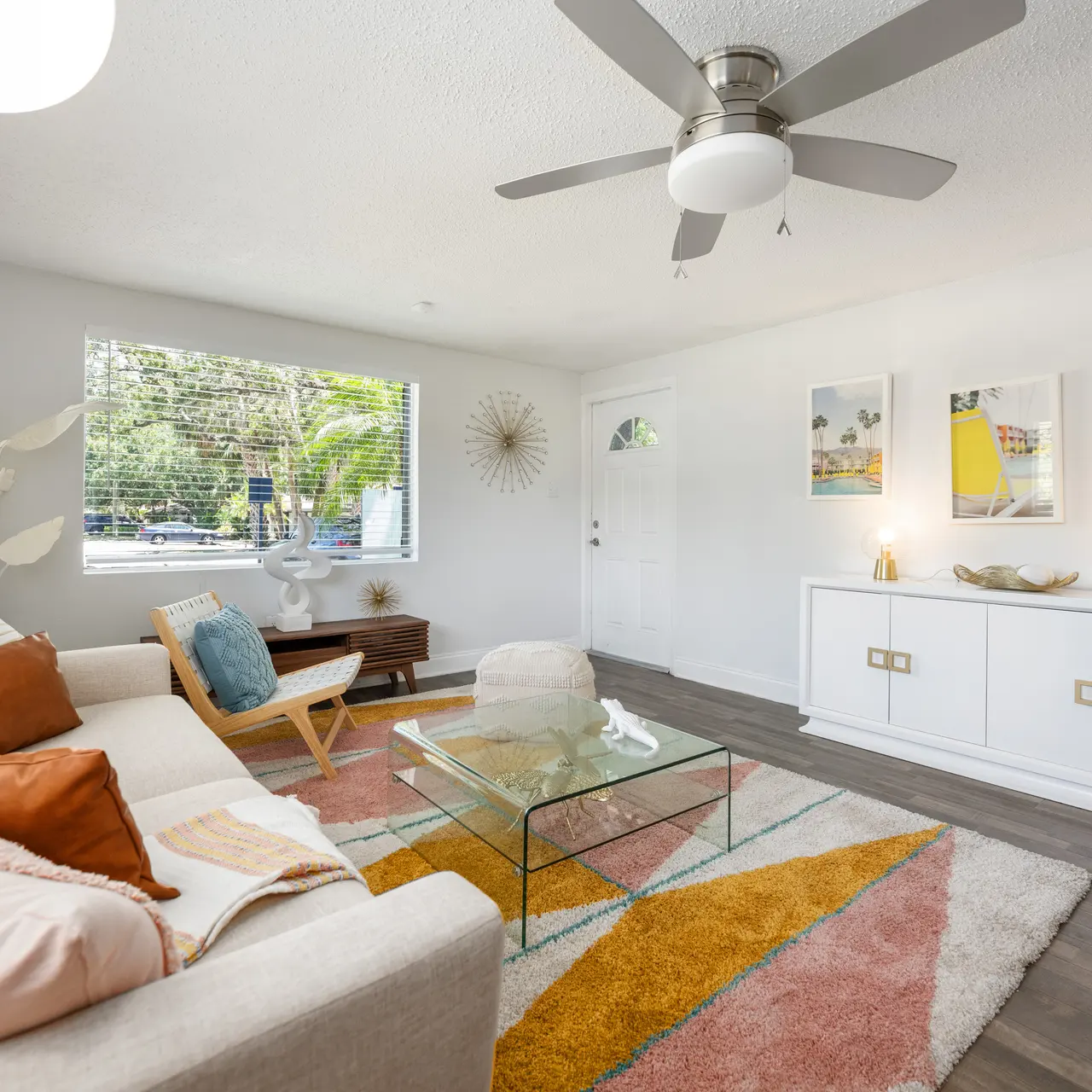 A bright and airy modern living room featuring a light beige sofa adorned with colorful cushions, a glass coffee table on a vibrant area rug, and a white media console. Large window allows natural light, and tasteful wall art adds character.