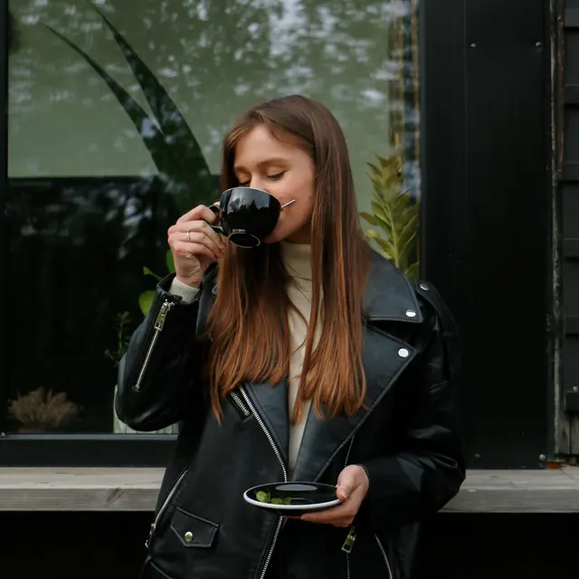 A young woman with long brown hair, wearing a black leather jacket and holding a black cup, stands next to a window with greenery visible outside.