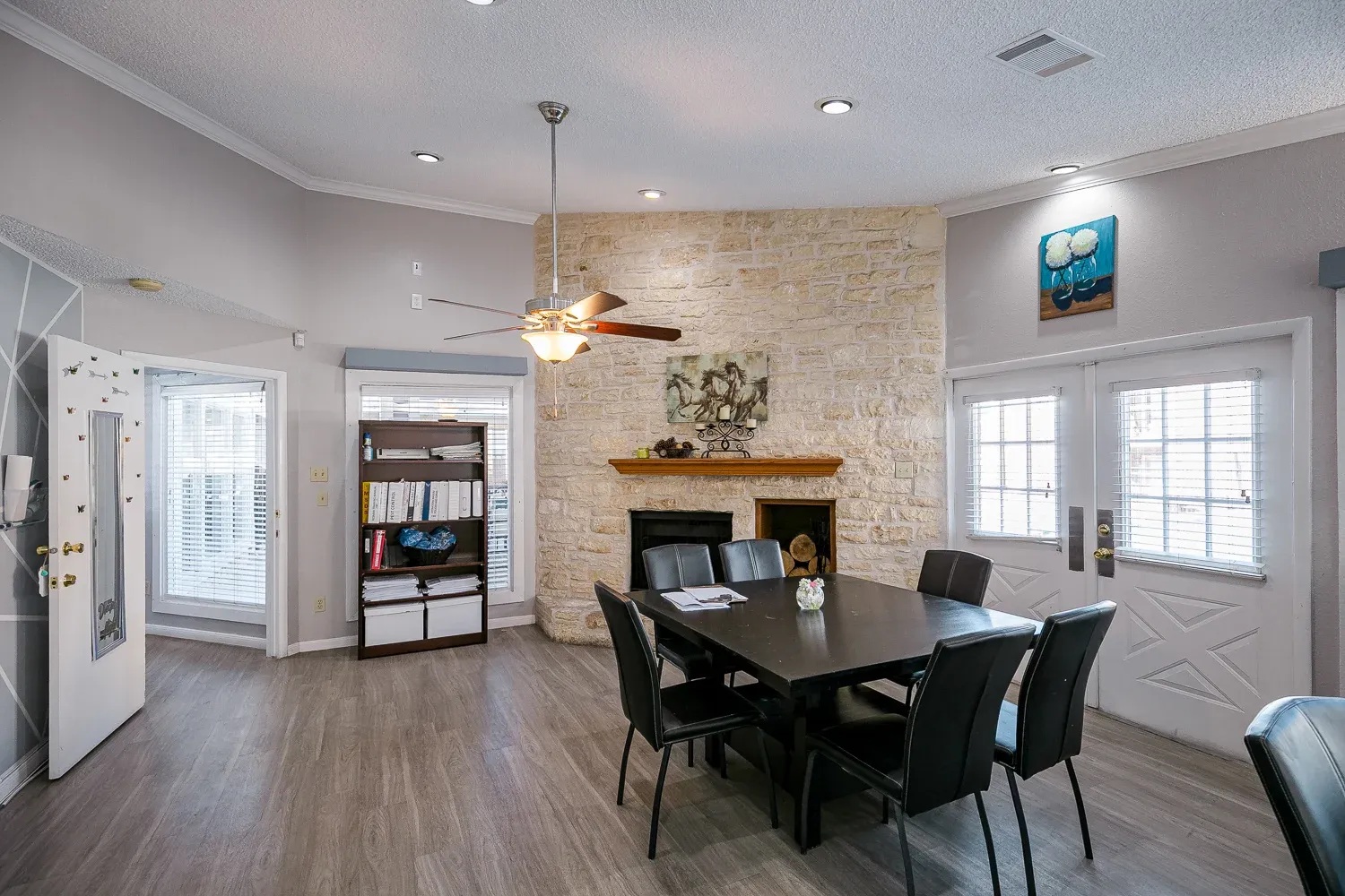 Stylish Dining Area with Stone Fireplace A modern dining area featuring a black table surrounded by black chairs, a stone fireplace, and large windows with natural light. Decorative wall art can be seen above the fireplace, and a ceiling fan is installed in the high ceiling.