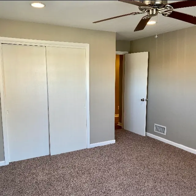 A spacious empty bedroom featuring beige walls, a ceiling fan, and carpeted flooring. There are two closet doors on the left and a doorway leading to another room on the right.