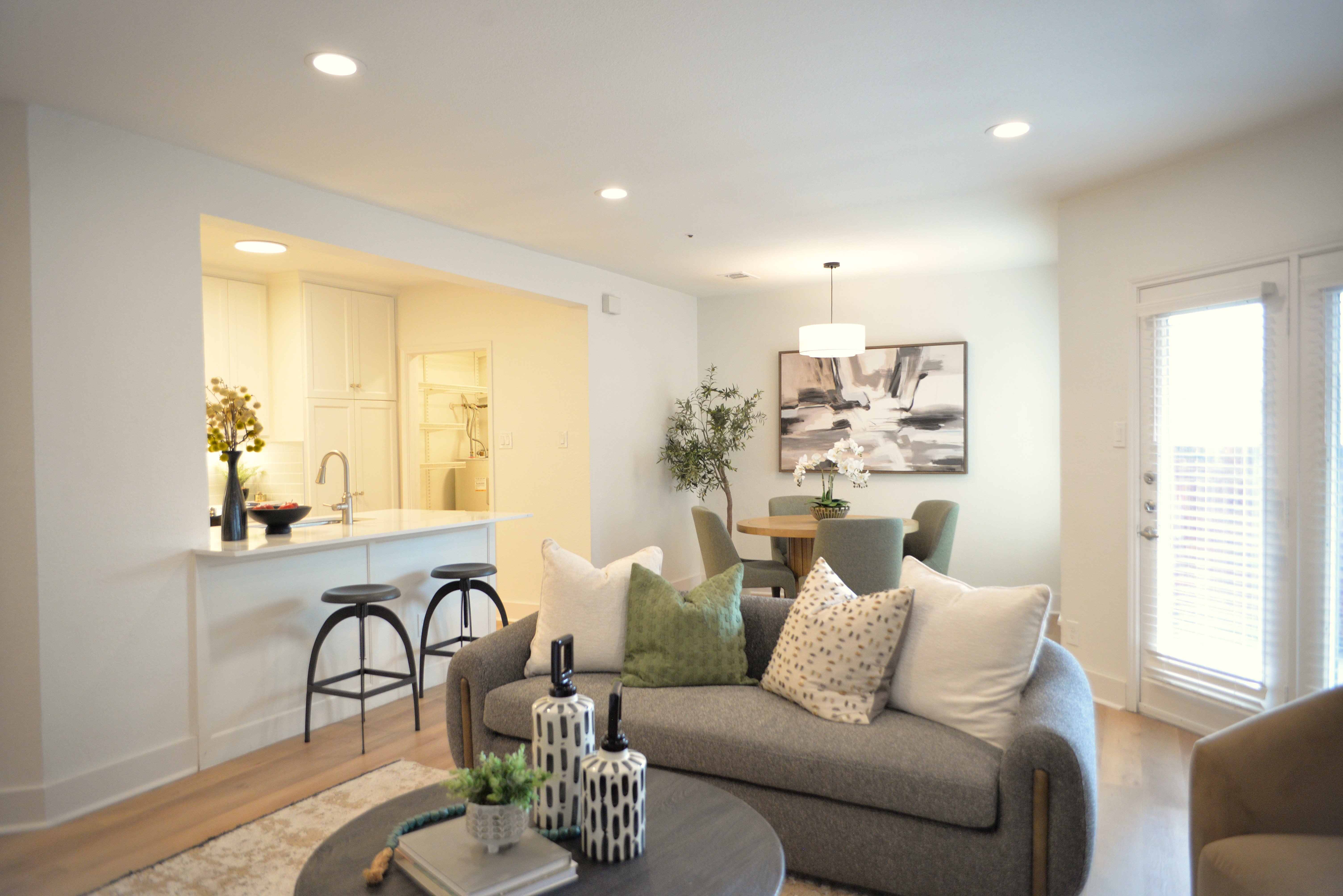 Contemporary Living Room and Dining Area A modern living room featuring a gray sofa adorned with decorative pillows, a round coffee table with decorative items, and a dining area with chairs in the background. The room has bright lighting and a minimalist design.