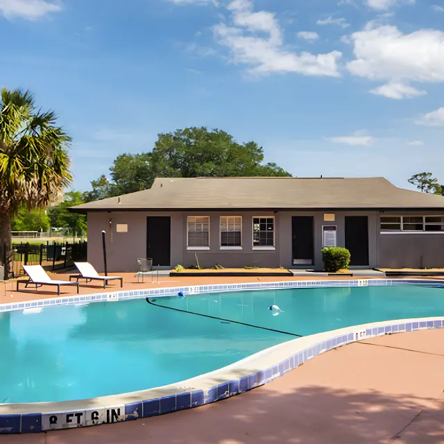 A bright outdoor swimming pool area with palm trees, lounge chairs, and a building in the background.