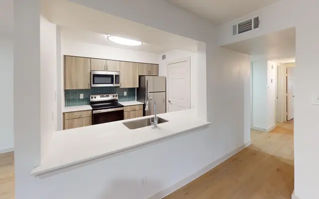 The Gleason A modern kitchen viewed from an open wall. The kitchen features wooden cabinets, stainless steel appliances, and a quartz countertop. There is a sink in the foreground and an entrance to a hallway in the background.