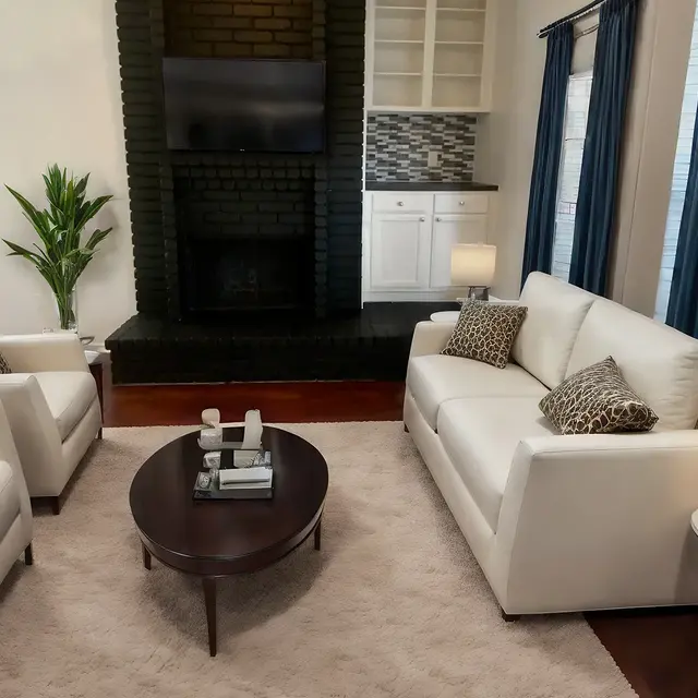A modern living room featuring two white sofas, a round coffee table, and a decorative plant. The room has large windows with blue curtains, a grey area rug, and a dark fireplace with a TV mounted above it.