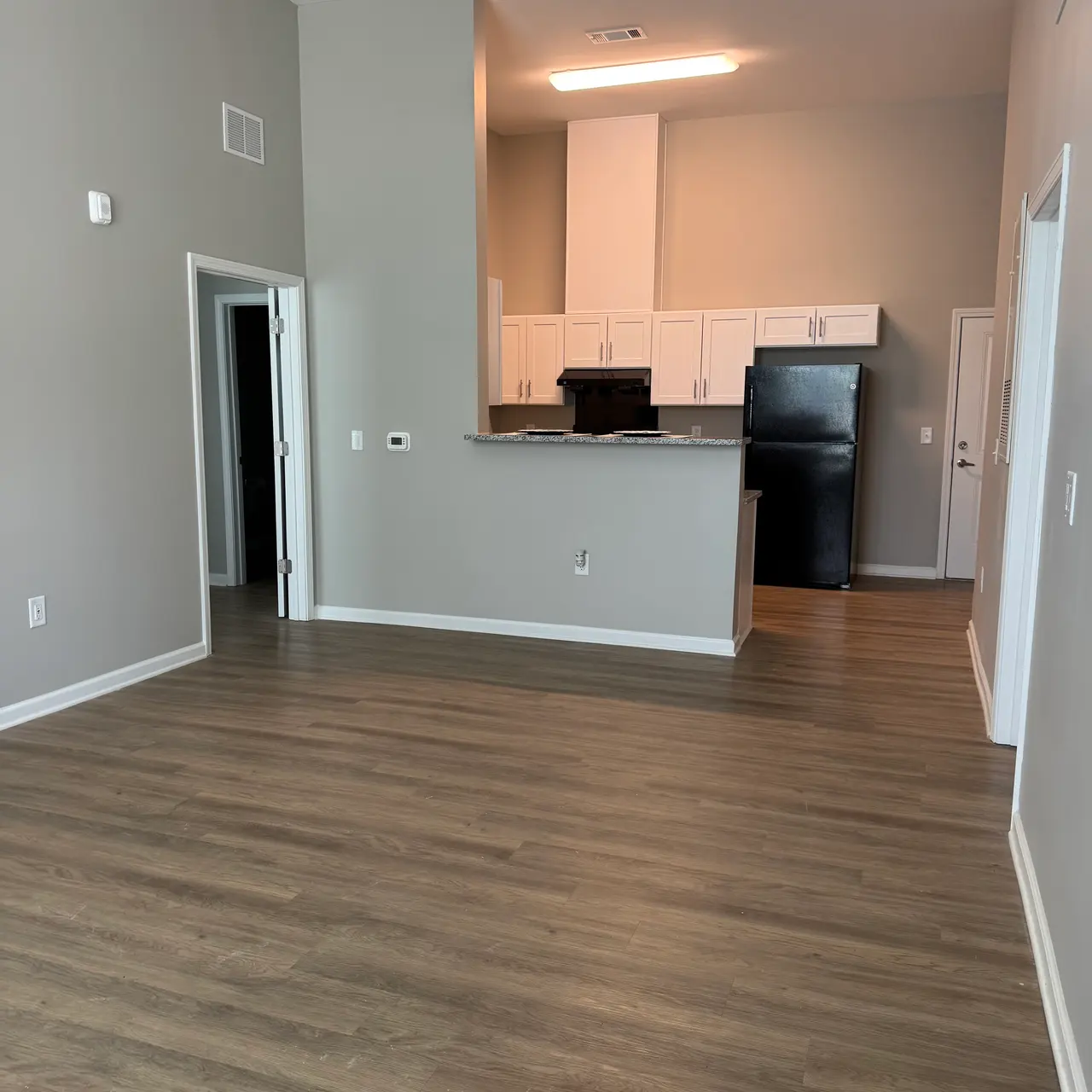 A spacious living area in a modern apartment featuring light wood flooring, a kitchen with white cabinets and black appliances, and neutral gray walls.