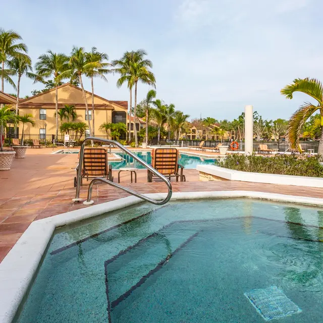 A scenic view of a swimming pool area featuring a hot tub, palm trees, and lounge chairs on a sunny day.